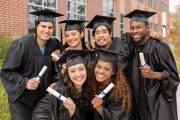A group of happy graduates in caps and gowns celebrate while holding their diplomas outside a brick building.